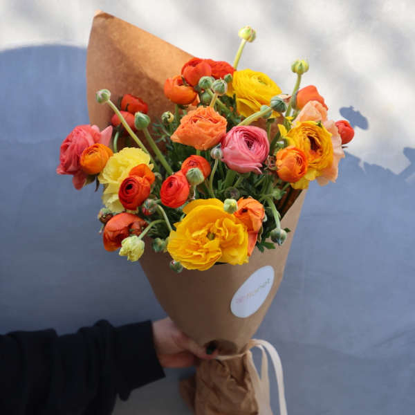 Handheld bouquet of orange, yellow, and pink ranunculus wrapped in brown paper
