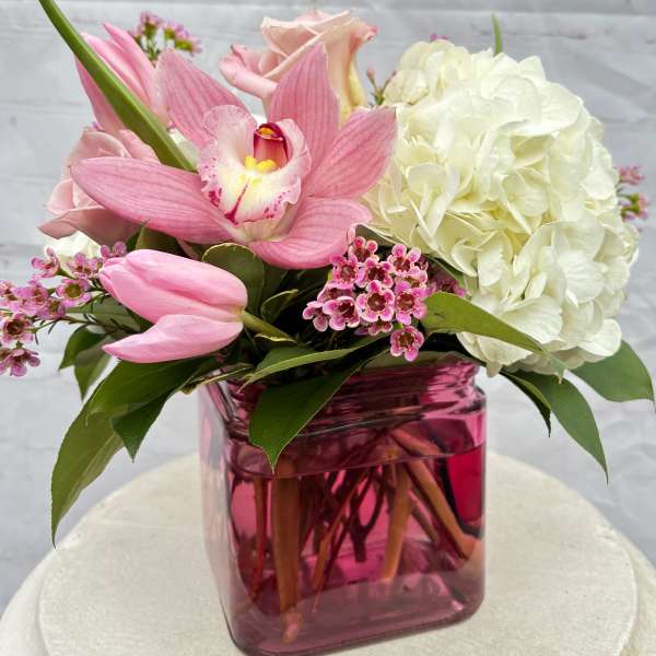 Low pink and white flower arrangement with orchid, tulips, roses, and hydrangea in a pink glass cube vase.