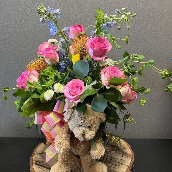 Pink roses and blue flowers arranged with a teddy bear and ribbon