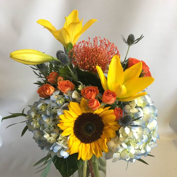 Bouquet of yellow lilies, sunflowers, orange roses, and blue hydrangeas in a glass vase