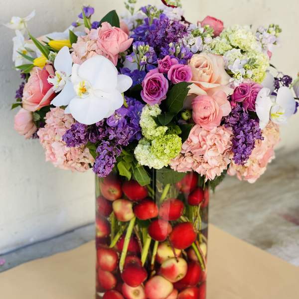 Mixed pink, purple, and white flowers in a tall glass vase filled with apples