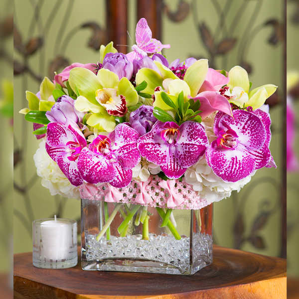 Pink orchids and lilies arranged in a square glass vase beside a white candle