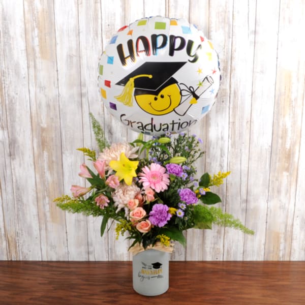 Graduation balloon with smiley face paired with mixed bouquet of pink, yellow, and purple flowers in a white vase.