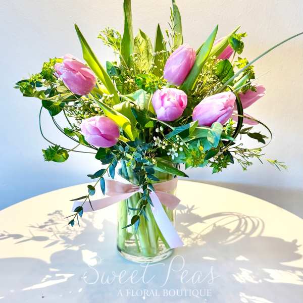 Pink tulips arranged in a clear glass vase with a pink ribbon on a white table.