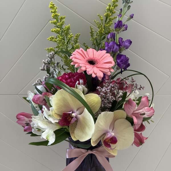 Tall arrangement of pink gerbera, orchids, and mixed flowers in a purple vase with a pink ribbon bow