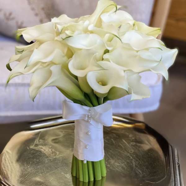 Hand-tied bouquet of white calla lilies wrapped in white ribbon with pearls, resting on a metallic tray