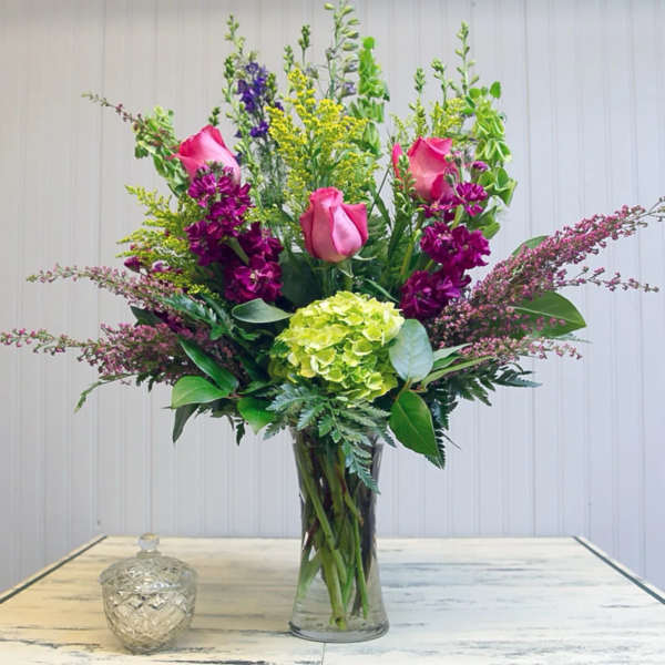 Tall bouquet of pink roses, purple blooms, and green hydrangea in a glass vase