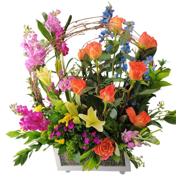 Tall mixed arrangement of orange roses, yellow lilies, and bright blooms in a white rectangular container