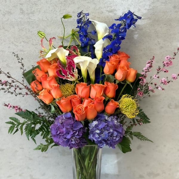 Tall mixed bouquet with orange roses, blue delphinium, white calla lilies, and purple hydrangeas in a glass vase