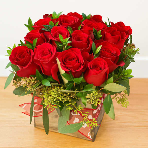 Red rose arrangement in a square glass vase with greenery and ribbon