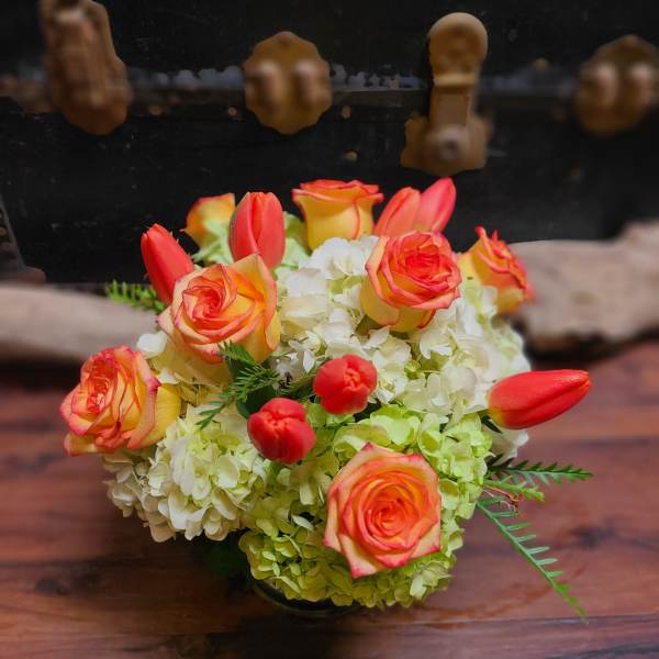 Low arrangement of orange roses and tulips with pale green hydrangeas in a glass vase