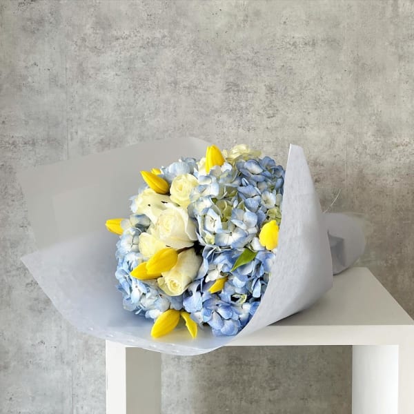 Bouquet of blue hydrangeas, white roses, and yellow tulips wrapped in white paper
