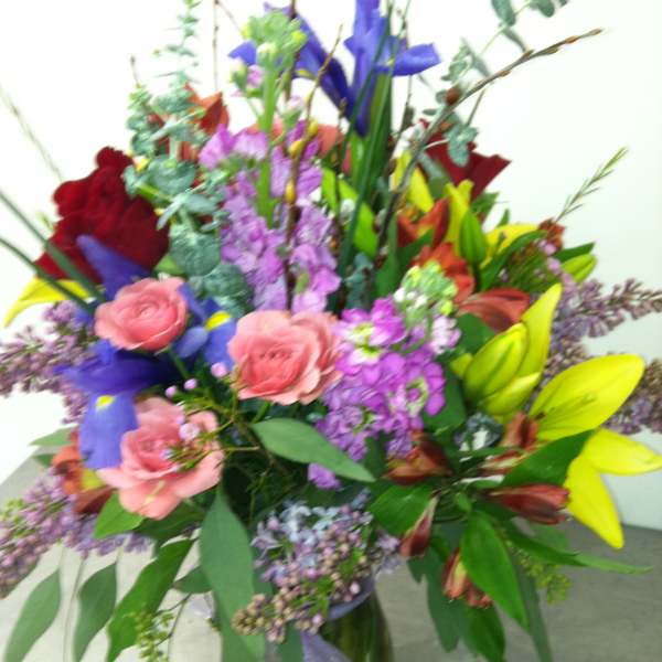 Mixed bouquet of pink roses, purple blooms, yellow lilies, and red flowers in a clear glass vase with ribbon