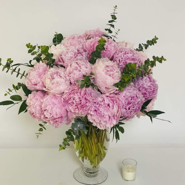 Large arrangement of pink peonies in a clear glass vase beside a small votive candle