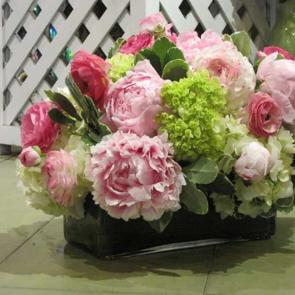 Low arrangement of pink peonies, ranunculus, and white and green hydrangeas in a rectangular glass vase