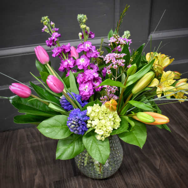 Mixed bouquet in a textured glass vase with pink tulips, purple blooms, and yellow lilies
