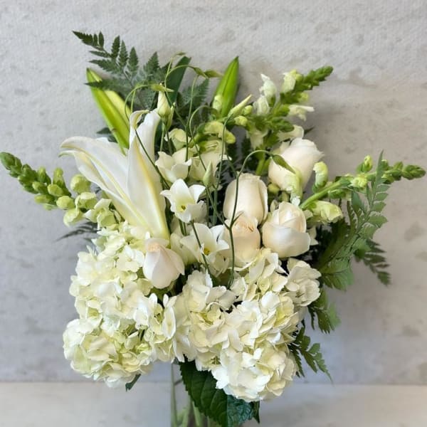 White floral arrangement in a glass vase with lilies, roses, and hydrangeas