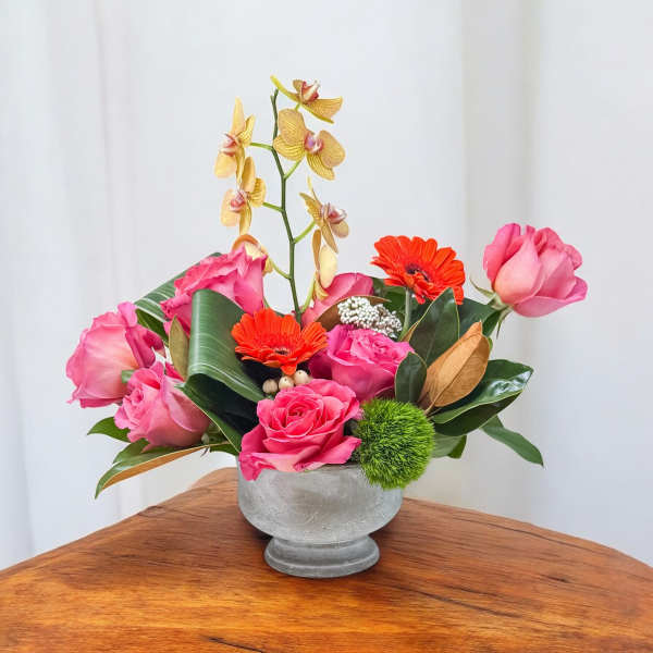 Pink roses and orange gerberas in a gray vase with a tall orchid stem