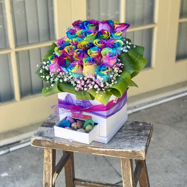Rainbow-colored roses arranged in a white box with purple ribbon and chocolates