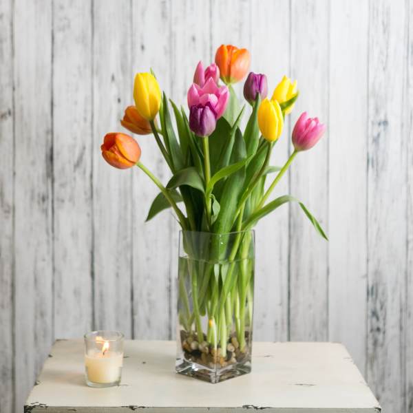 Tall mixed-color tulips in a clear glass vase with pebbles beside a lit votive candle.