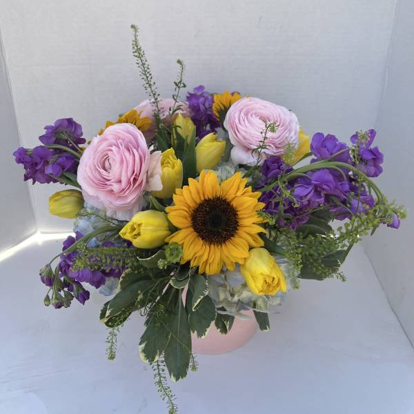Bouquet with pink ranunculus, yellow tulips, purple flowers, and a sunflower in a pink container
