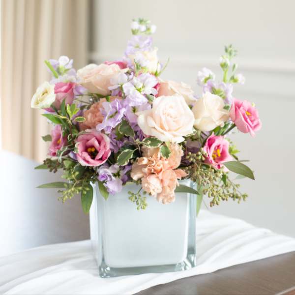 Low pastel arrangement of roses and lisianthus in a frosted glass cube vase on a draped table
