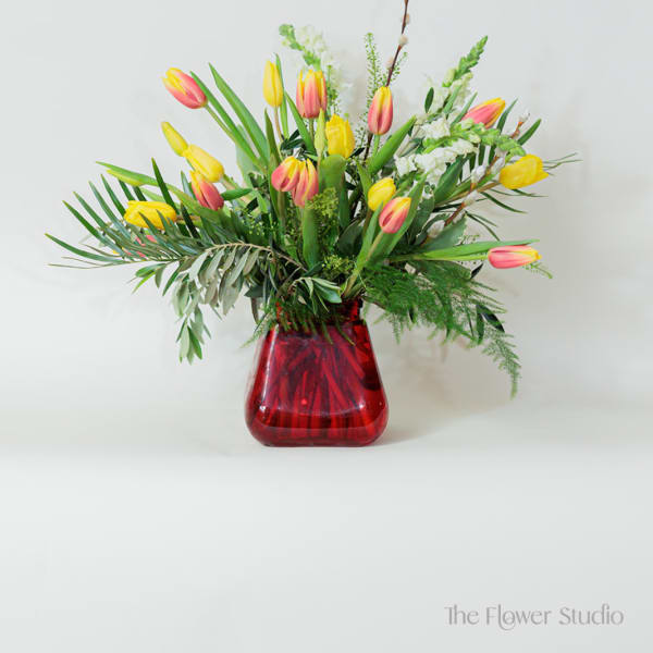 Yellow and pink tulips with white snapdragons and greenery in a red glass vase