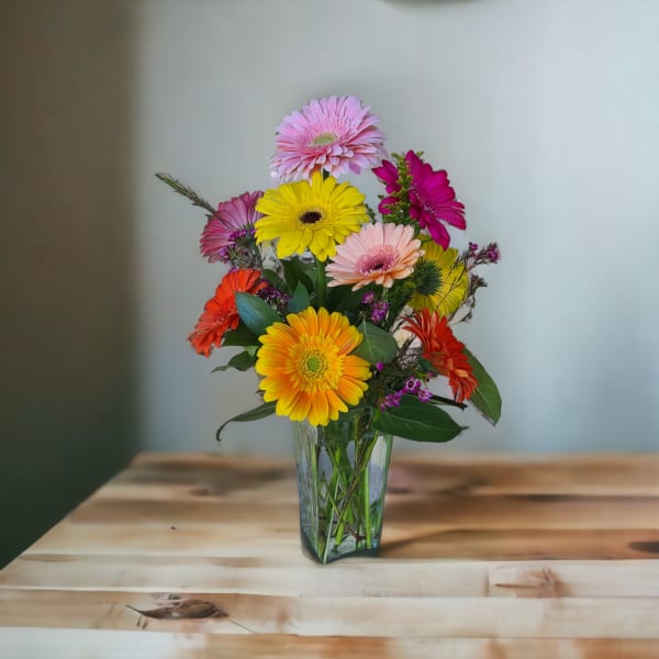 Tall arrangement of colorful gerbera daisies in a clear glass vase on a wooden table.