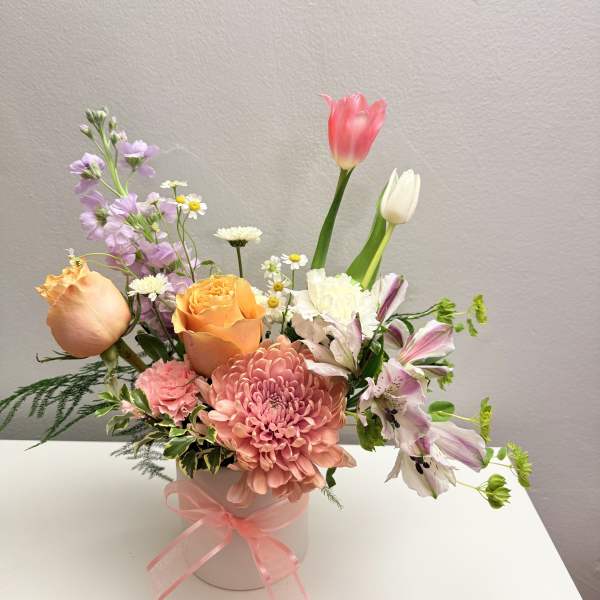 Pastel arrangement with peach roses, pink chrysanthemum, tulips, and mixed blooms in a white vase with pink ribbon