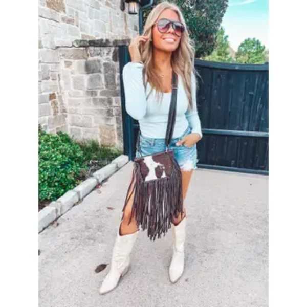Woman wearing sunglasses, a light top, denim shorts, and white boots with a fringe crossbody bag.