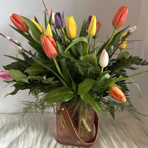 Mixed-color tulip arrangement in a pink glass handled vase with lush foliage.
