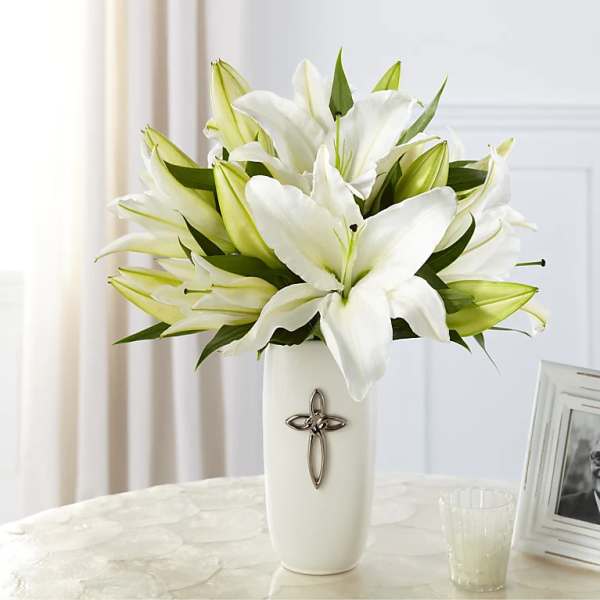 White lilies arranged in a tall white vase with a silver cross emblem on a light table