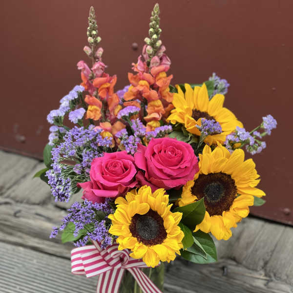 Bouquet of pink roses, sunflowers, and purple filler in a glass vase with a striped ribbon.