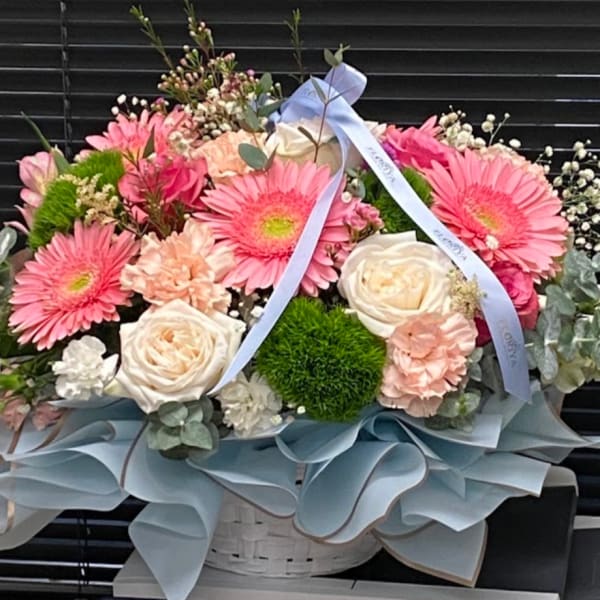 Pink gerbera daisies and cream roses arranged in a basket with blue ribbon