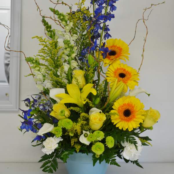 Tall mixed bouquet in a blue pot with yellow gerberas, blue delphinium, and white lilies