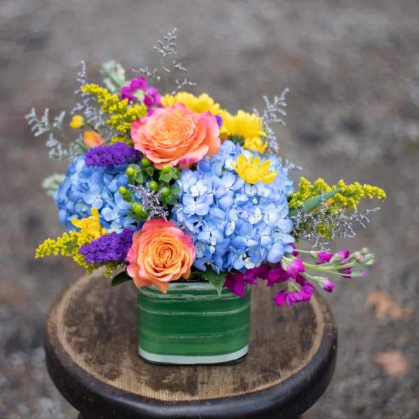 Colorful bouquet in a green square vase with blue hydrangeas and orange roses