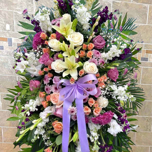Tall standing spray of white lilies, roses, and purple flowers with a lavender bow on an easel.