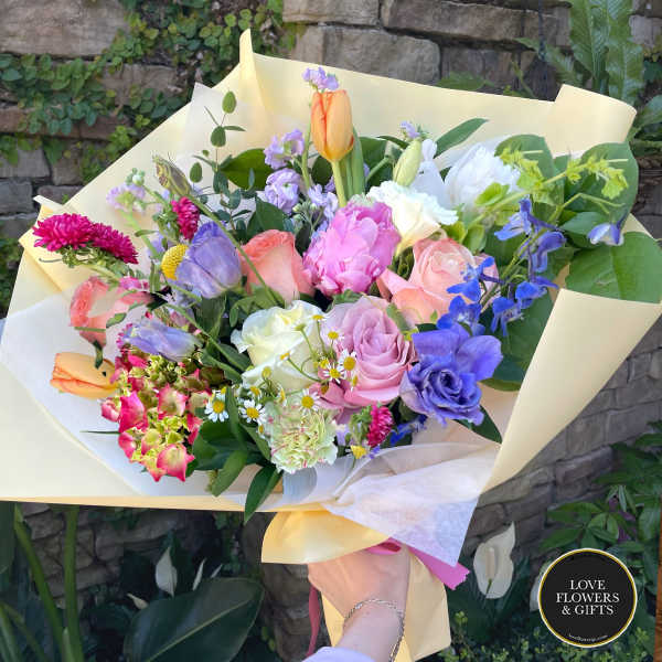Hand-tied bouquet of mixed pastel roses, tulips, and other flowers wrapped in pale yellow paper