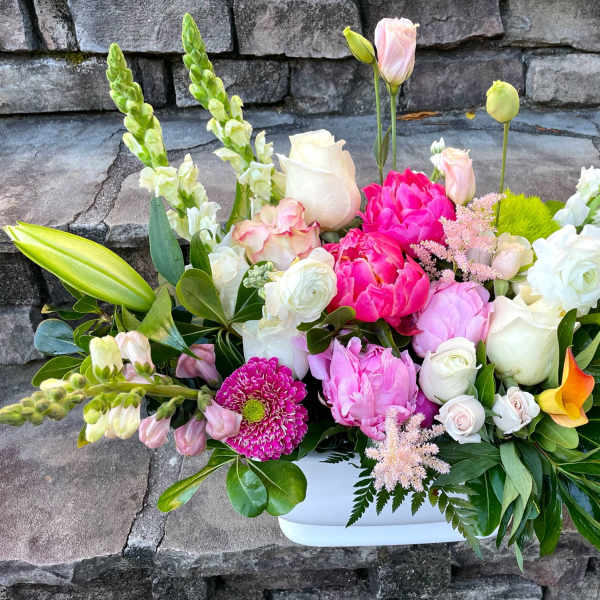 Rectangular arrangement of pink peonies, white roses, snapdragons and orange calla lilies in a white container.
