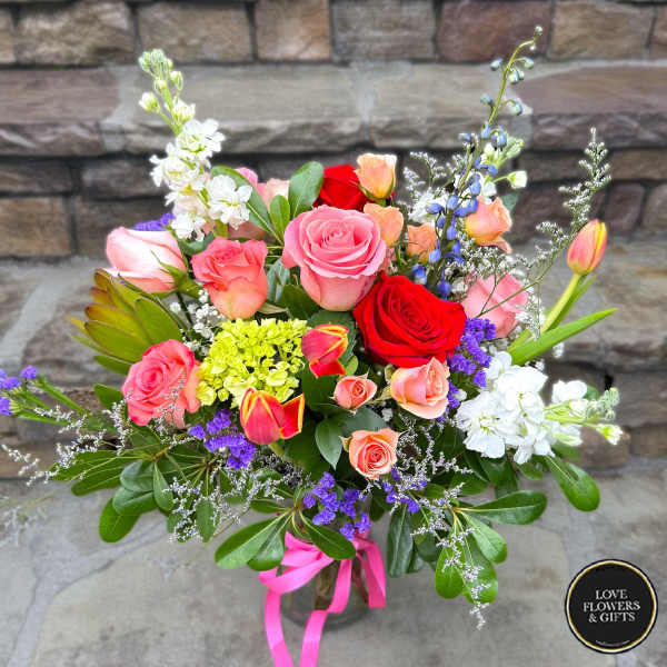Mixed bouquet of pink and red roses, tulips, and other flowers in a glass vase with a pink ribbon