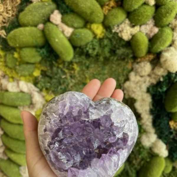 Hand holding a heart-shaped purple crystal against a green floral wall