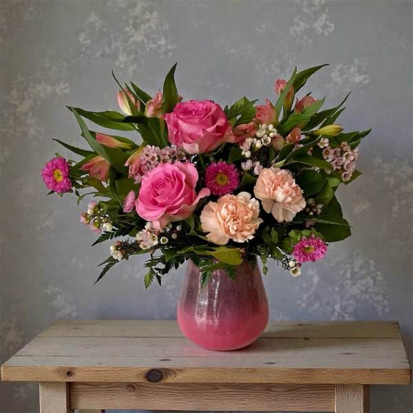 Pink and peach flower arrangement with roses and carnations in a pink glass vase on a wooden table