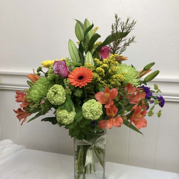 Tall mixed bouquet of orange, green, yellow and pink flowers in a clear rectangular glass vase with ribbon