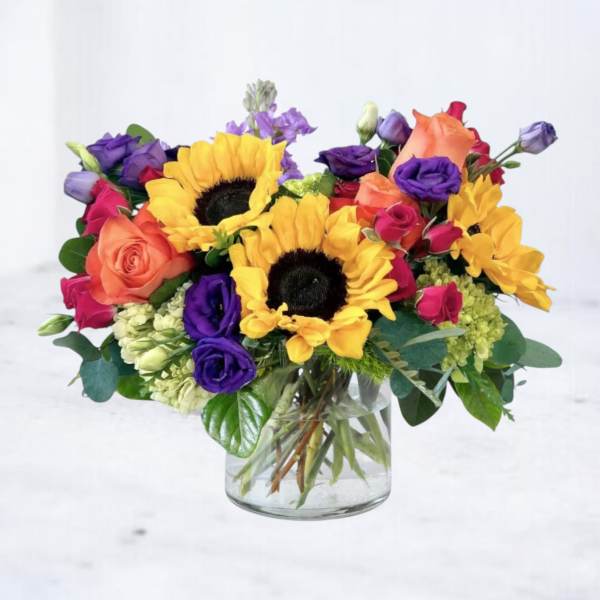 Bright arrangement of sunflowers, roses, and purple blooms in a clear glass vase
