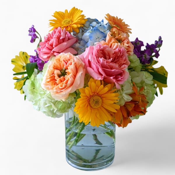 Mixed bouquet of yellow gerbera daisies, pink and peach roses, and hydrangeas in a clear glass vase