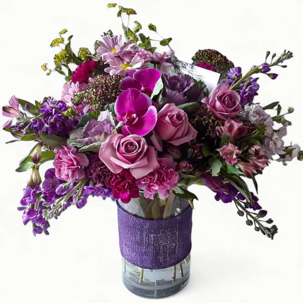 Purple and pink mixed flower arrangement with roses and orchids in a clear vase wrapped with purple fabric