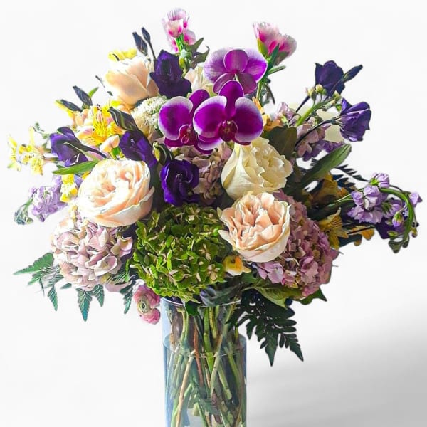 Tall glass vase of mixed pink roses, hydrangeas, and purple orchids with assorted accent flowers.