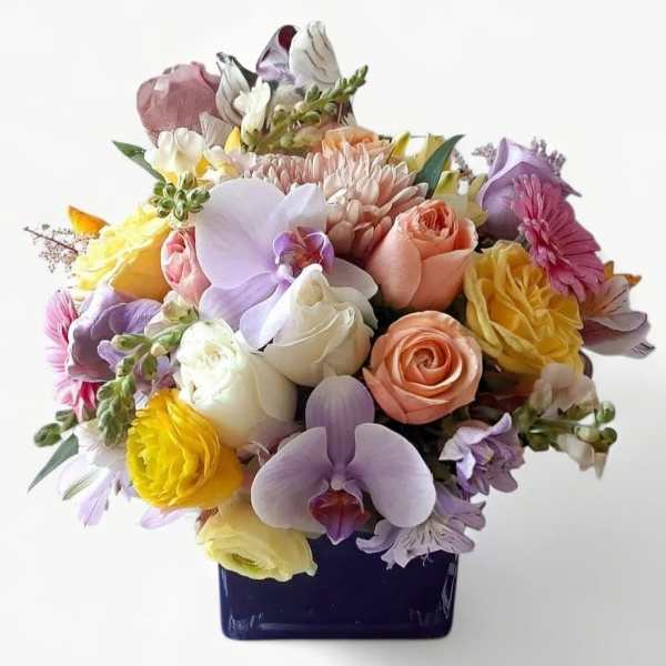 Pastel mixed flower arrangement with roses, orchids, and daisies in a dark blue square vase