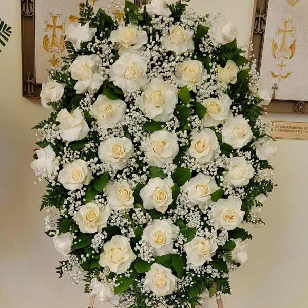 Oval standing spray of white roses and baby's breath on an easel with a black dedication sign.