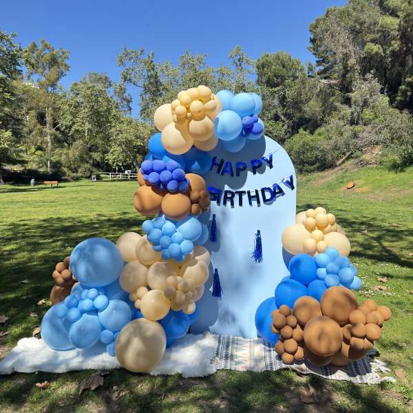Outdoor birthday backdrop with blue arch and clusters of blue, tan, and brown balloons on a lawn
